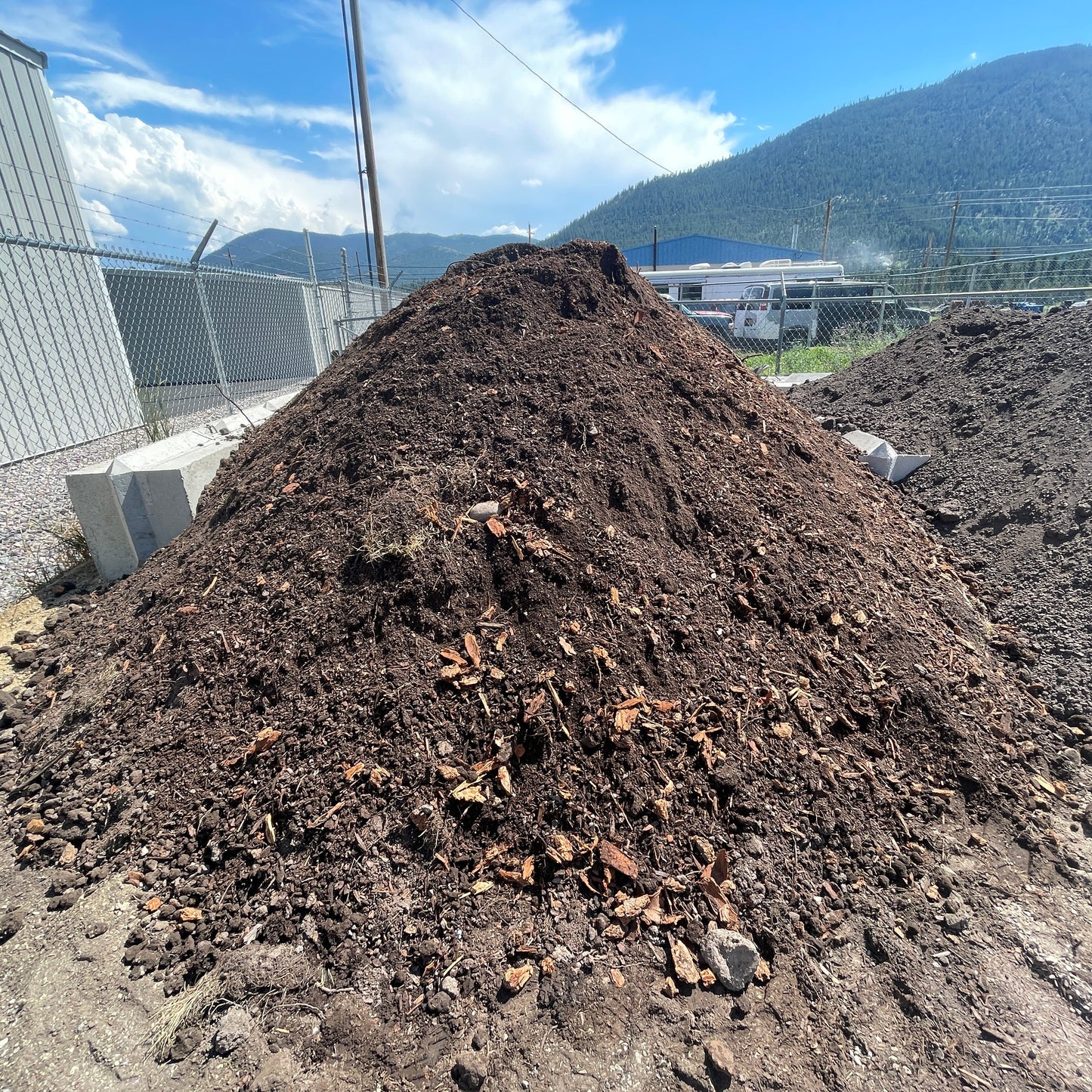 A large mound of dark brown unscreened  soil sits outdoors beside a chain-link fence, with mountains, blue sky, and buildings visible in the background.